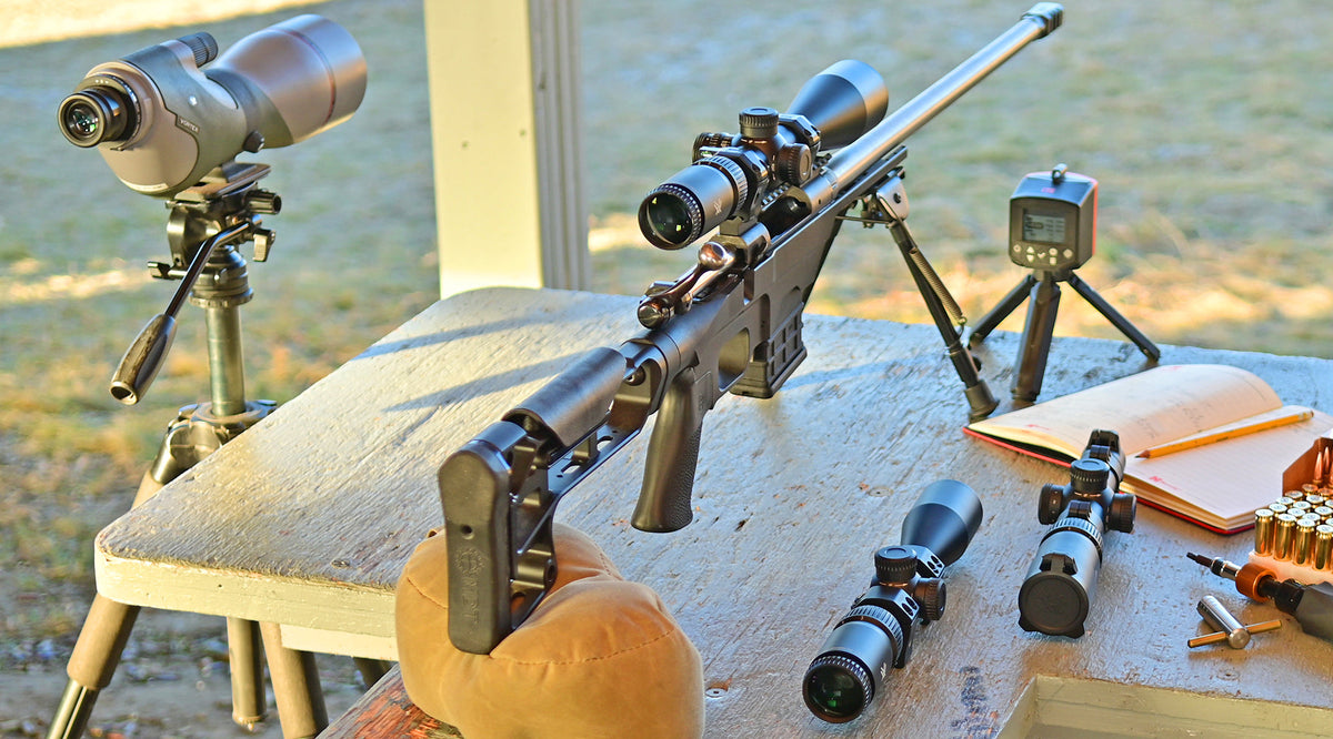 Bolt-action rifle in a black MDT chassis on a bipod at a shooting bench with a spotting scope, chronograph, spare scopes, torque driver and ammunition